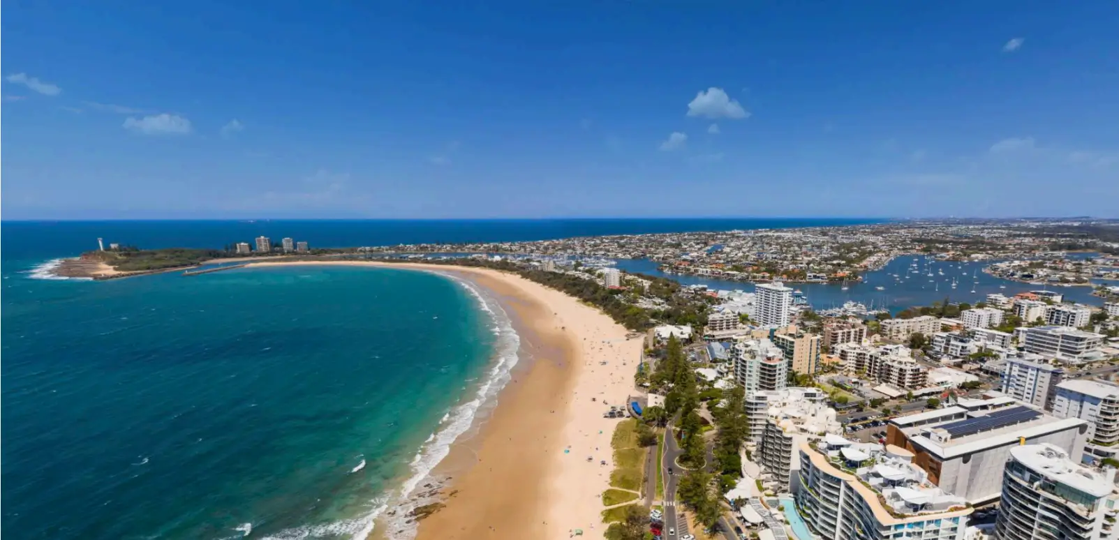 Aerial view of a curved sandy beach along a turquoise bay, with a dense city of high-rise buildings along the shore and a marina on the right.
